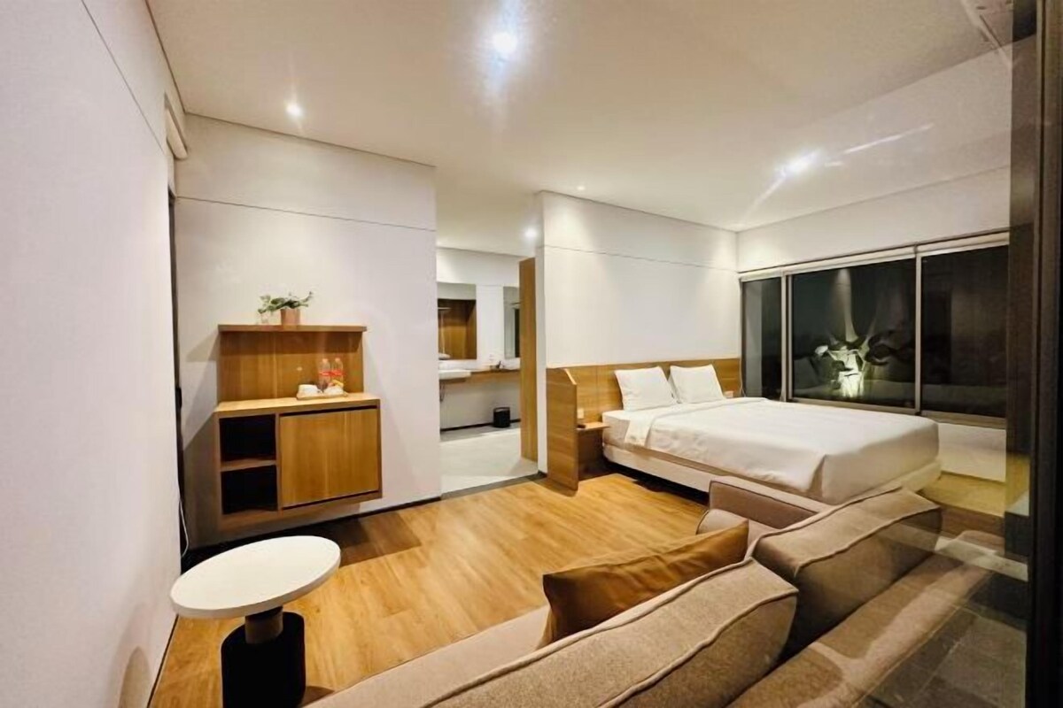 A modern hotel room with a bed, a small desk, a seating area, and a large window.