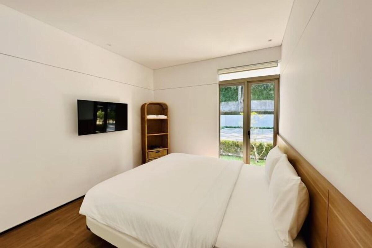 Une chambre moderne avec un grand lit, un téléviseur fixé au mur, une étagère en bois et une fenêtre donnant sur un paysage verdoyant.