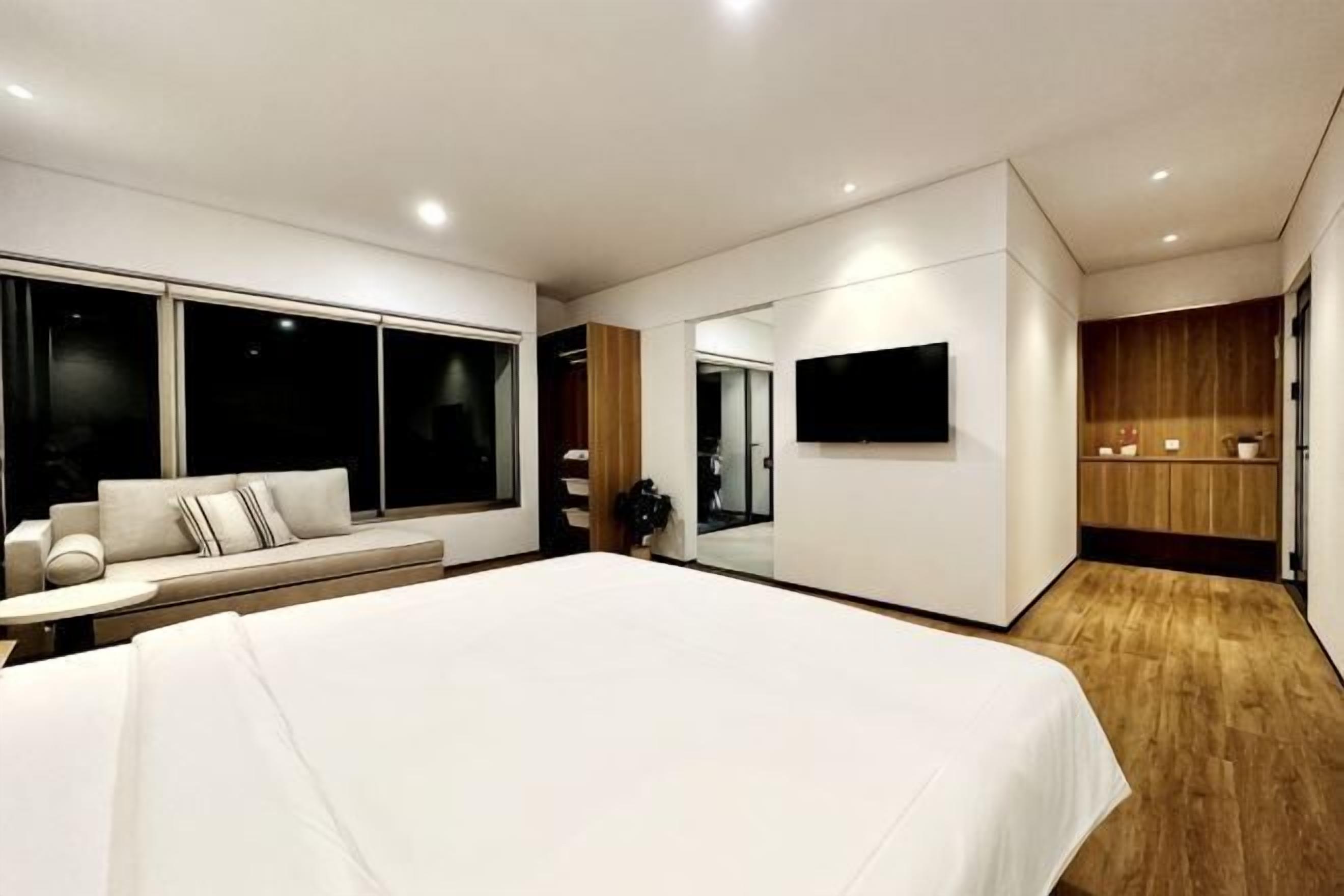 A modern bedroom with a large bed, a seating area with a sofa, a TV mounted on the wall, and a wooden floor.