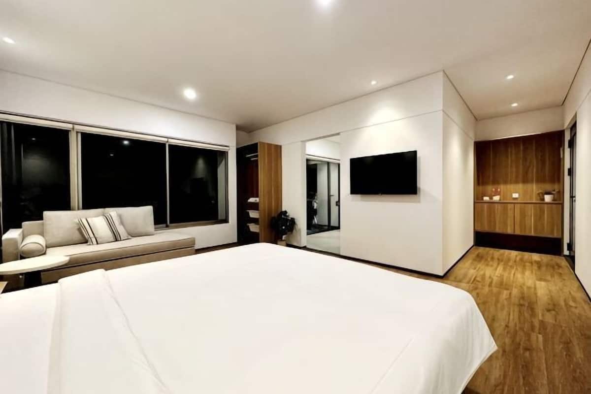 A modern bedroom with a large bed, a seating area with a sofa, a TV mounted on the wall, and a wooden floor.