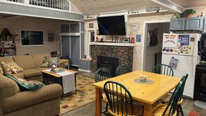 Una sala de estar acogedora con un sofá, una mesa de centro y una mesa de comedor con sillas. Hay una chimenea, un televisor y un refrigerador con imanes.