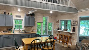 Cocina rústica y acogedora con comedor, pisos de madera y ventanas amplias que dan a la vegetación exterior.