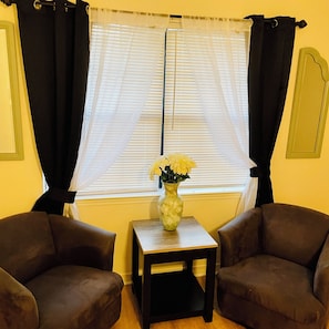 Ein Zimmer mit zwei braunen Sesseln, einem kleinen Holztisch mit einer Vase voller gelber Blumen und einem Fenster mit weißen Rollos und schwarzen Vorhängen.