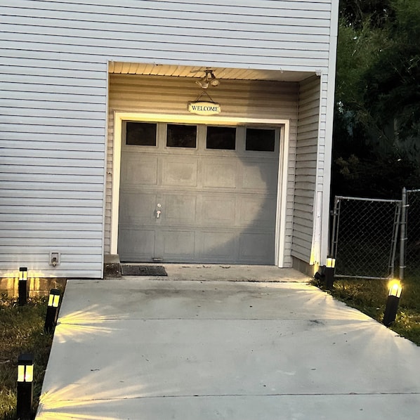 Eine Garage mit einem 'WELCOME'-Schild über der Tür, einer Betonauffahrt und einem Zaun aus Maschendraht zur rechten Seite.