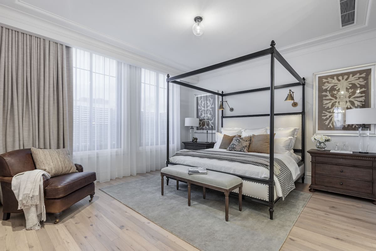 A four-poster bed with a bedside table and lamp, a leather armchair, a bench, and a dresser with a mirror.