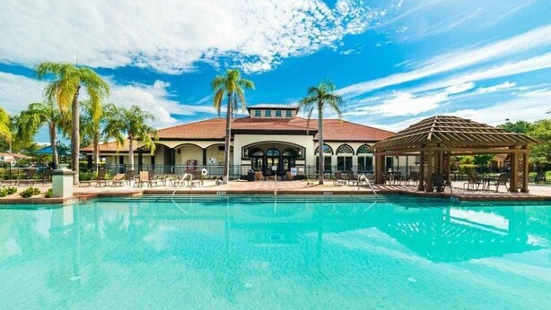 Zona de piscina con un edificio al fondo, palmeras y un pabellón.