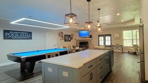 A modern living room with a pool table, a fireplace, and a kitchen area with a refrigerator. - Modern Farmhouse in Wallisville! (Wallisville)