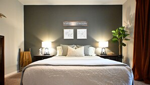 A neatly made bed with a white comforter and pillows, a dark wall with a quote, two framed pictures, and a plant on a nightstand. - Modern Farmhouse in Wallisville! (Wallisville)
