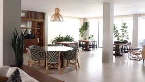 Un comedor amplio con mesas y sillas redondas, rodeado de ventanas grandes y plantas en macetas.