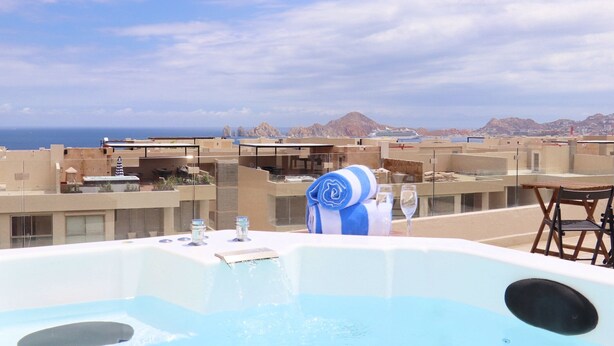 Zona de piscina en la azotea con un jacuzzi, una mesa con una copa de vino y una silla. La vista incluye un paisaje montañoso y un cielo despejado.
