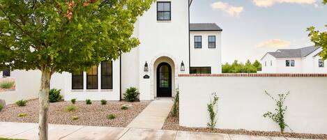 A modern two-story house with a white exterior, a front entrance with an archway, and a small front yard with a tree and shrubs.