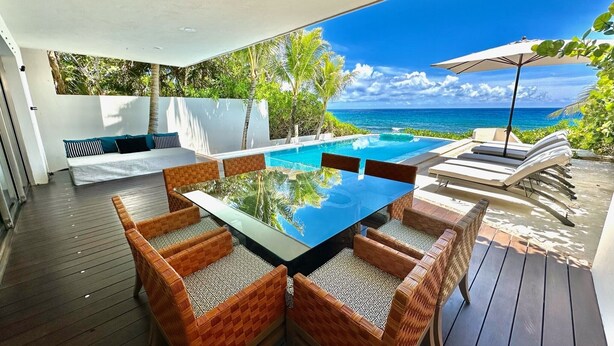 Un área de comedor al aire libre con una mesa de vidrio y sillas de mimbre, con vista a una piscina y al océano.