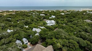 An aerial view of a residential area with houses surrounded by dense greenery, a beach in the background, and a clear sky. - Sundays at the Millers - BOTH CLUBS (Bald Head Island)