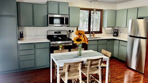 A kitchen with green cabinets, a stainless steel refrigerator, a microwave, and a stove. There is a dining table with chairs and a vase with sunflowers. - Relax at the Cozy Cabin, just a 5-min drive to it all (Harbor Springs)