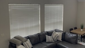 A modern living room with a sectional sofa, large windows with blinds, a dining area, and a view of a mountainous landscape.