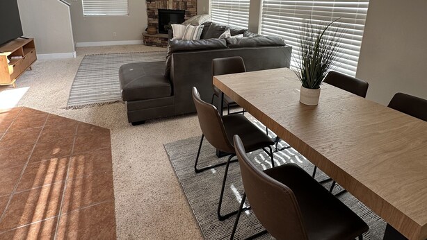 A modern living room with a stone fireplace, a dining area with a wooden table and chairs, and a large window with blinds.