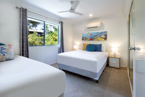 Quarto com duas camas, um ventilador de teto, um aparelho de ar condicionado e uma janela com cortinas.