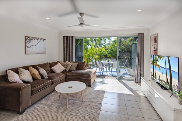 Relax in this stylish living room with a sofa, coffee table, and TV, offering a view of lush greenery and the beach.