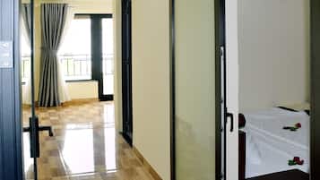 Una habitación de hotel con piso a cuadros, una cama con ropa blanca y rosas rojas, una puerta con picaporte y un ventanal con cortinas.