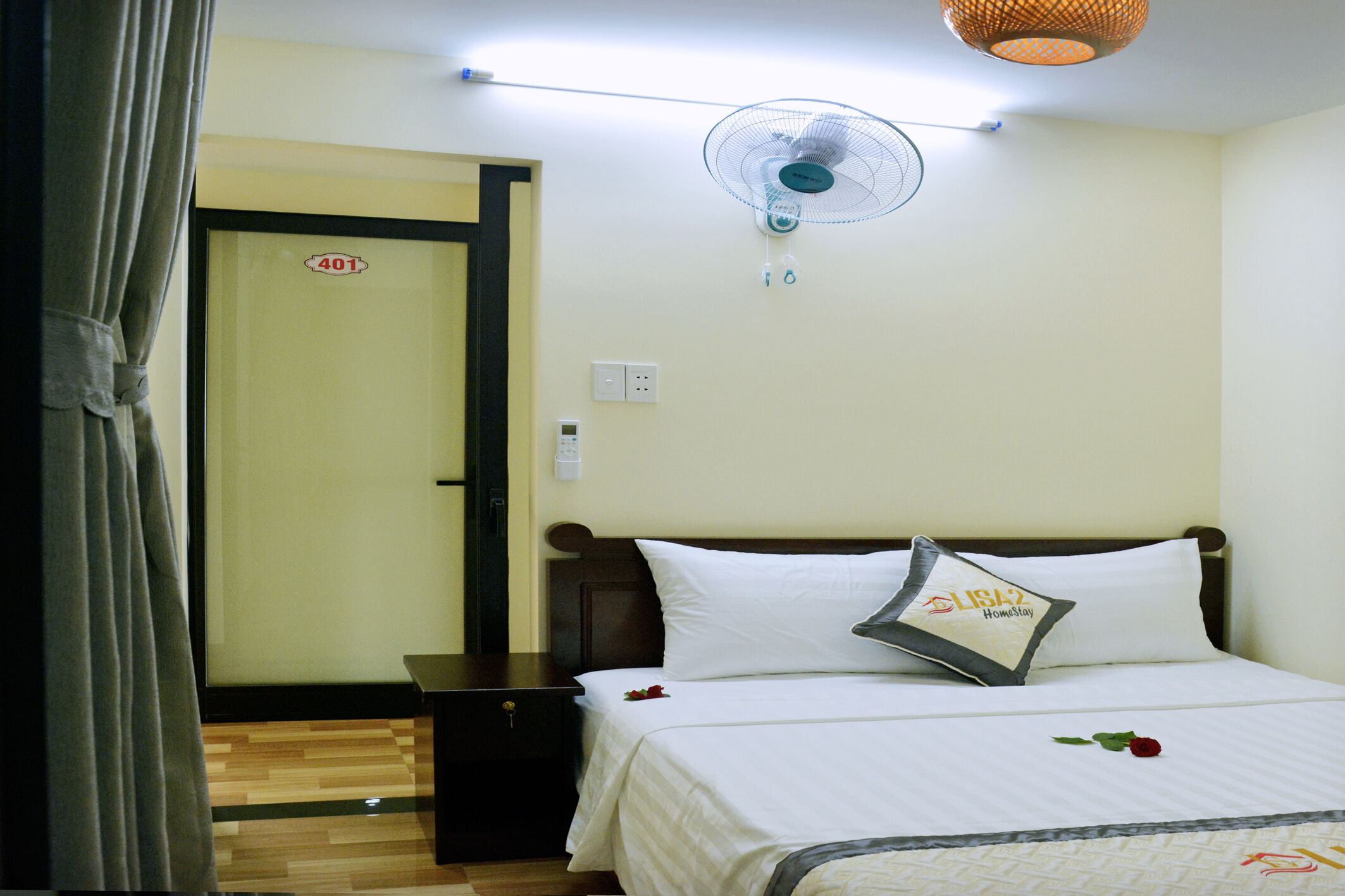 A hotel room with a bed, a bedside table, a nightstand, a wall-mounted fan, and a door with a number sign.