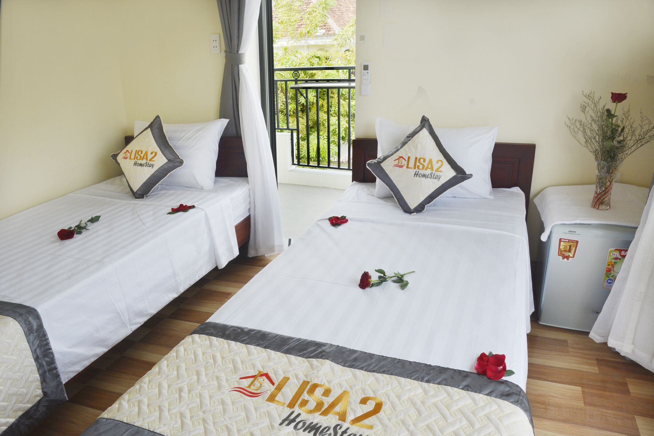 Une chambre d’hôtel avec deux lits, chacun recouvert d’une courtepointe blanche et agrémenté d’un oreiller orné du logo de l’hôtel. Des pétales de rose sont disposés sur les lits, et un réfrigérateur est visible dans un coin de la pièce.