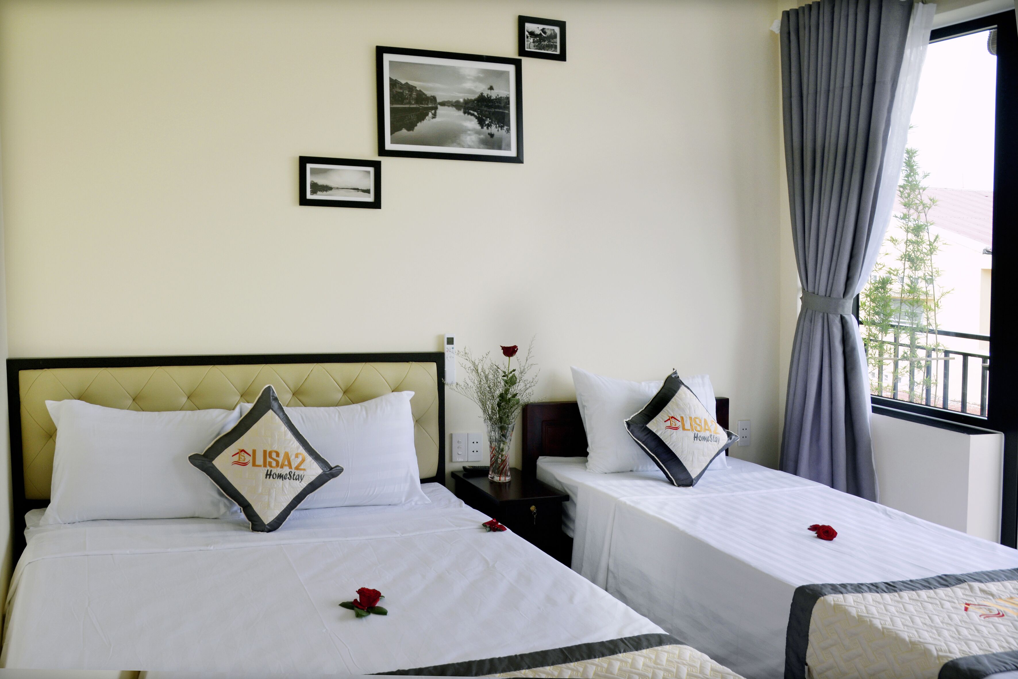 A hotel room with two beds, each with a pillow that reads "LISA2 HomeStay." There are framed pictures on the wall, a vase with flowers on the nightstand, and a window with curtains.