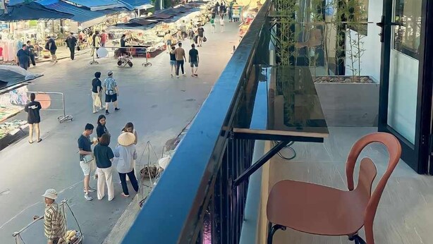 A balcony with a view of a bustling street market, a modern building with large windows, and a traditional building in the background. - Lisa Homestay 2 (Da Nang)