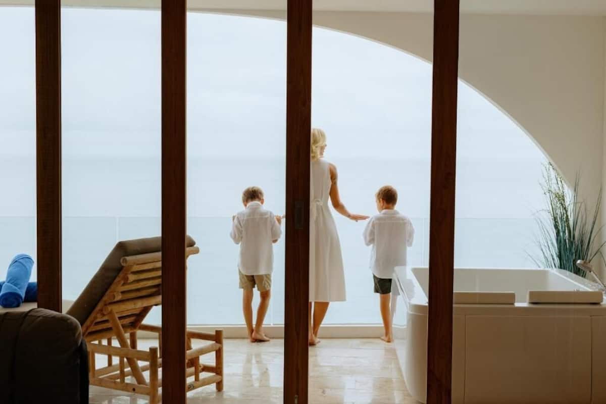 A room with a large arched window overlooking the sea, a wooden chair, and a bathtub.
