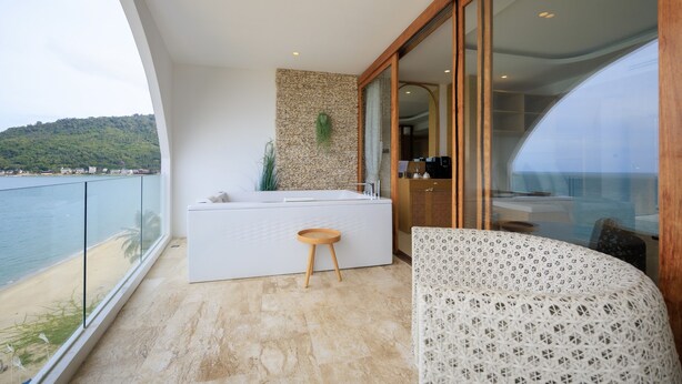 Un balcon avec vue sur la mer, une baignoire blanche et un fauteuil à motifs.