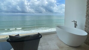 A modern bathroom with a freestanding bathtub and a wicker chair, overlooking a beach with waves and a cloudy sky. - Elite Atoll Beachfront Hotel (Khanom)