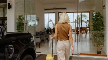 Eine Frau geht auf ein modernes Gebäude mit Glastüren zu. Ein geparktes Auto und ein gelber Koffer sind ebenfalls zu sehen.
