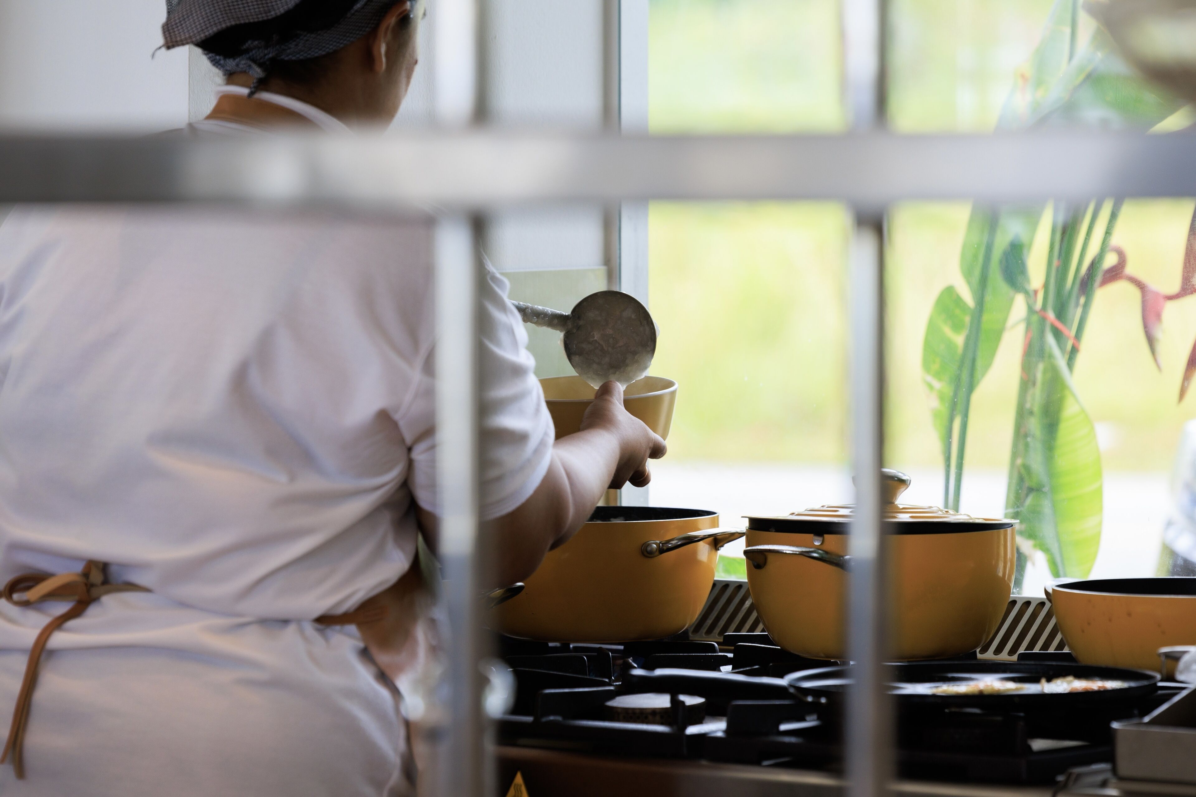A person cooking with pots on a stove, a window with a view of greenery, and a plant in the background.