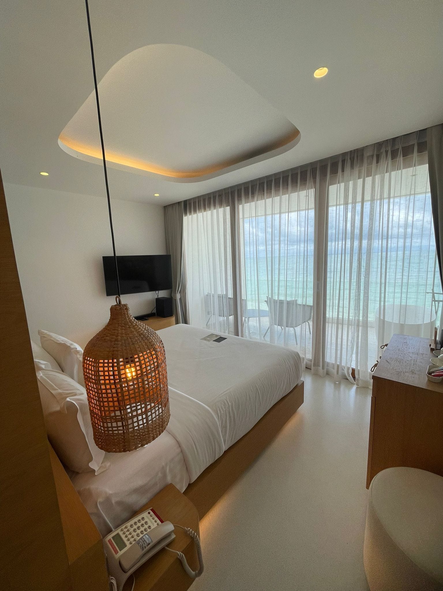 A modern hotel room with a large bed, a TV, a bedside table with a phone, and a view of the sea through sheer curtains.