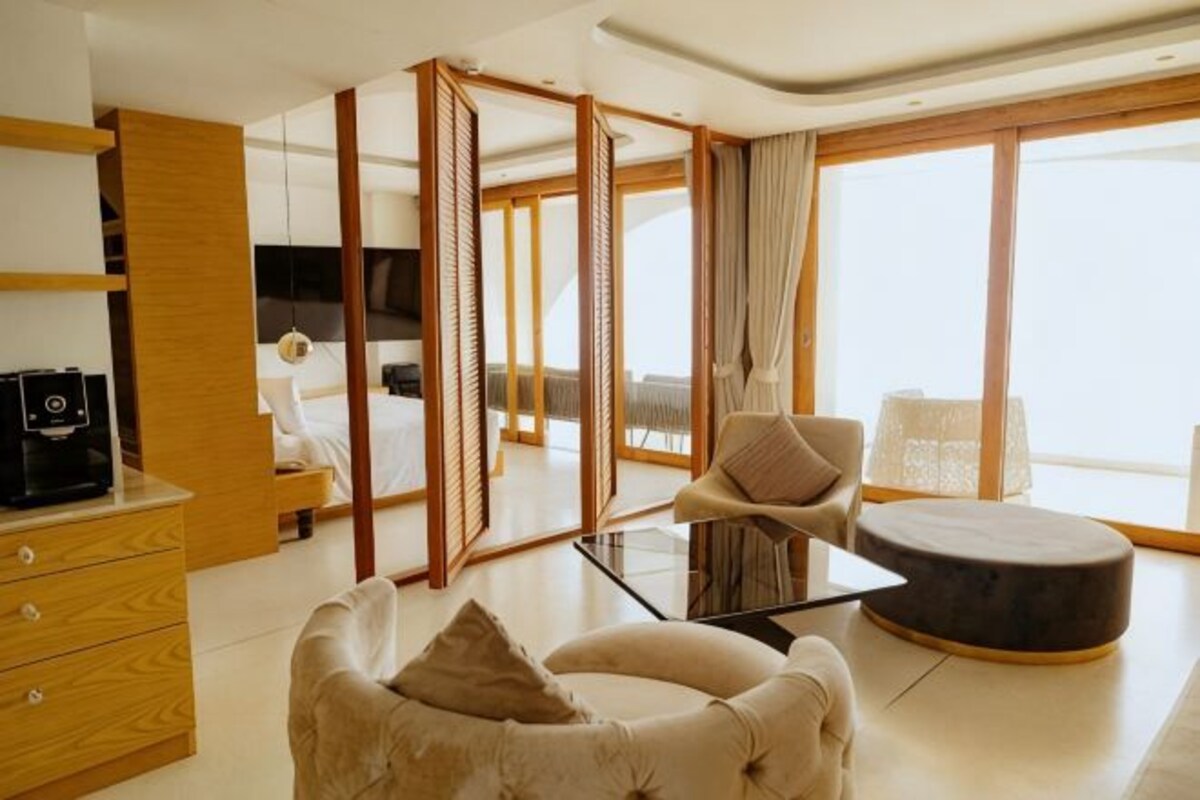 A modern hotel room with a sofa, a round ottoman, a TV, a desk with a computer, and a large window offering a view.