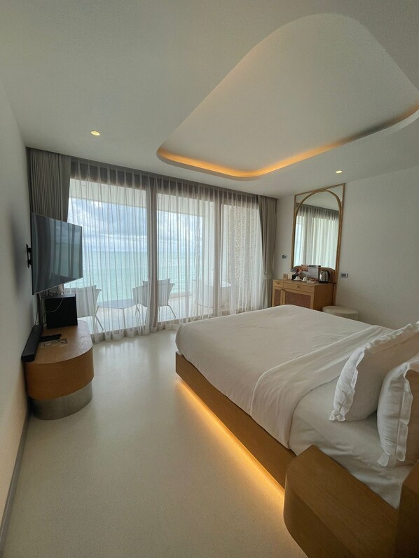 A modern hotel room with a large bed, a desk, a TV, and a view of the sea through sheer curtains.
