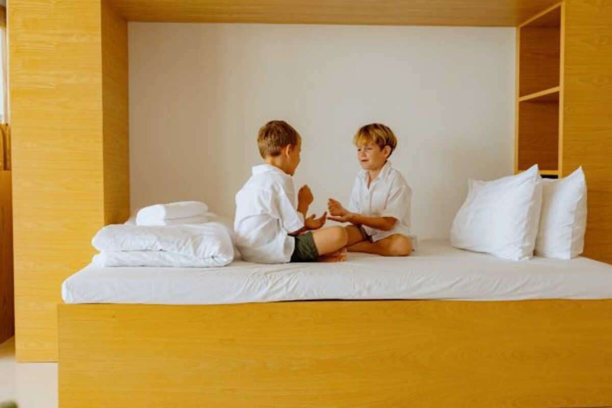 Twee kinderen zitten op een bed in een kamer met houten wanden en wit beddengoed.