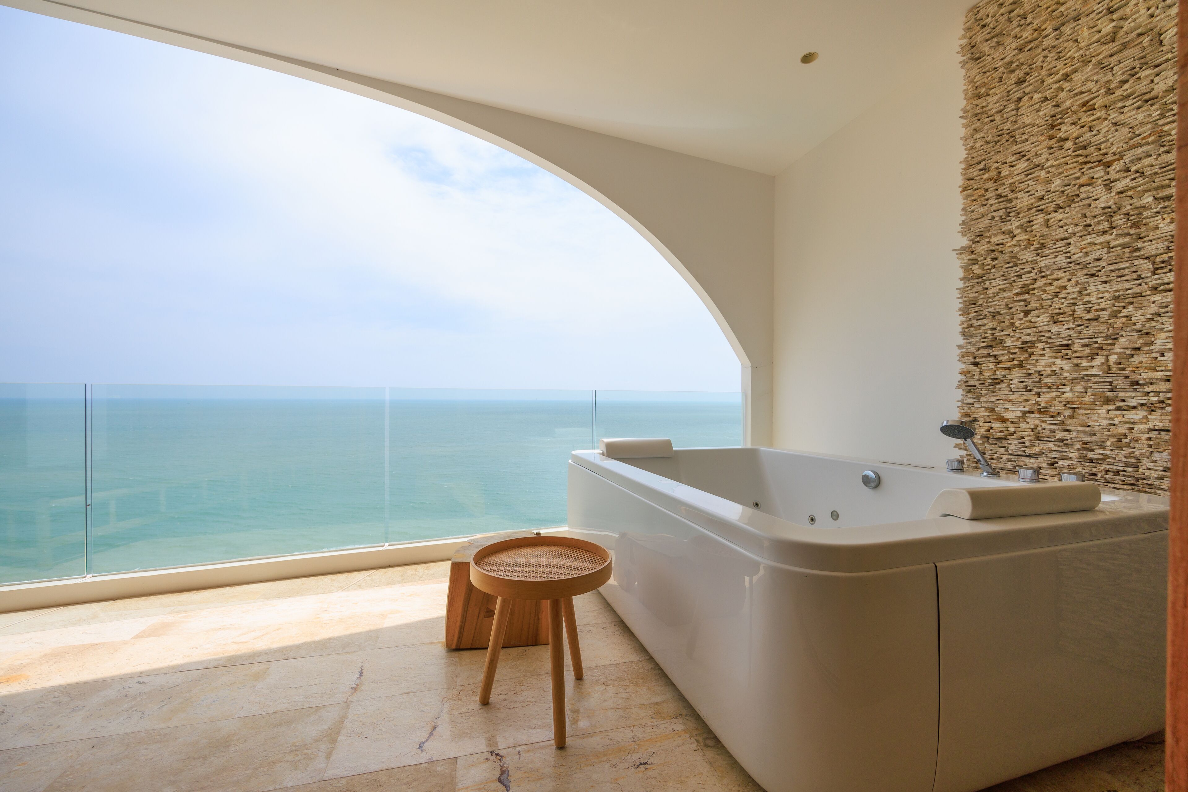 Ein modernes Badezimmer mit einer großen Badewanne, einem hölzernen Hocker und Meerblick.