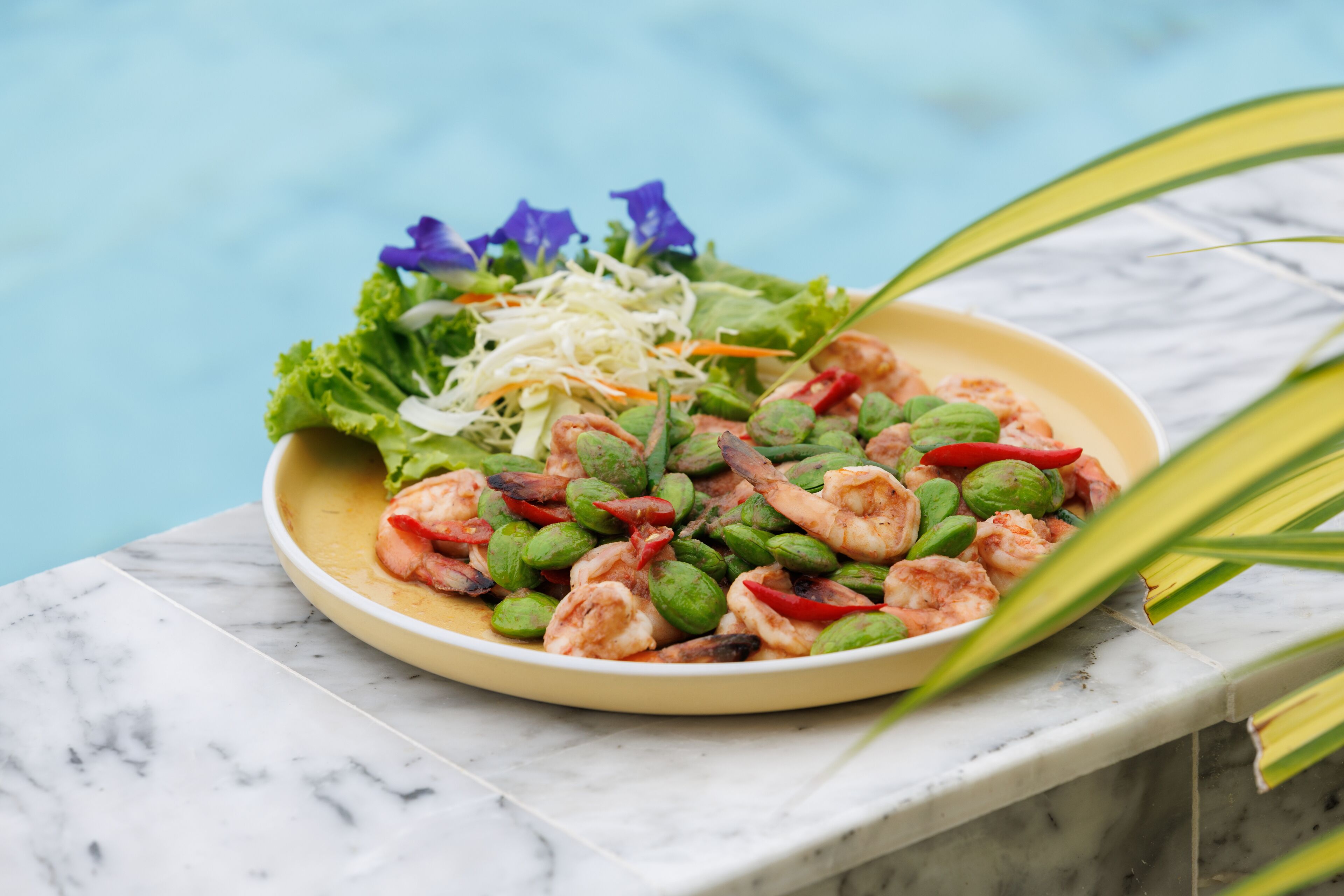 A plate of shrimp and vegetables on a marble surface near a pool.