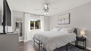 A bedroom with a bed, a ceiling fan, a nightstand with a lamp, a TV, and a window with curtains.