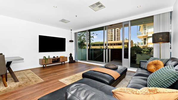 A modern living room with a large flat-screen TV, a black leather sofa, and a dining area with a wooden table and chairs.