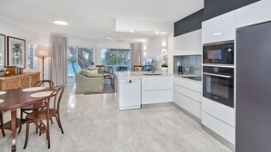 A modern kitchen with white cabinets, a Bosch oven, and a dishwasher. There is a dining area with a wooden table and chairs, and a view of the sea through large windows. - Coral Horizons Beachfront Apartment (Cairns)