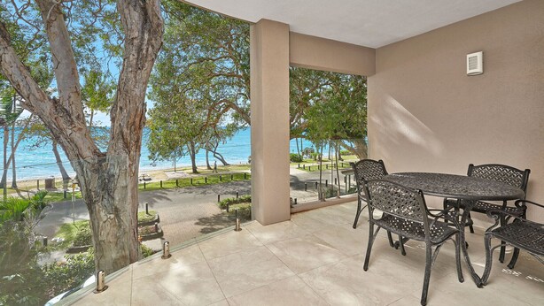 A covered patio with a table and chairs, overlooking a beach and trees. - Coral Horizons Beachfront Apartment (Cairns)