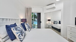 A hotel room with a large bed, a bedside table, a lamp, a dresser, and an air conditioning unit. - Coral Horizons Beachfront Apartment (Cairns)