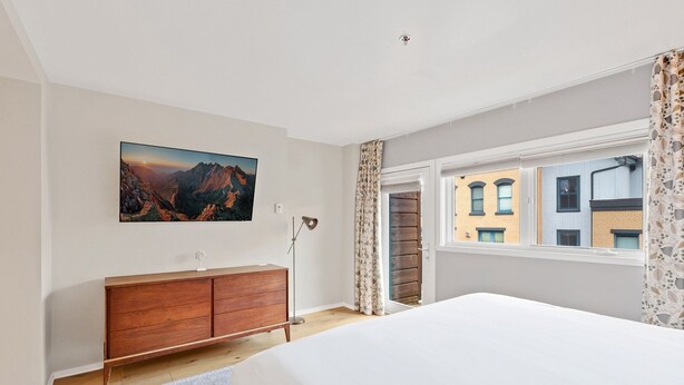 A bedroom with a large bed, a wooden dresser, a painting of a mountain landscape, and a window with a view of buildings.