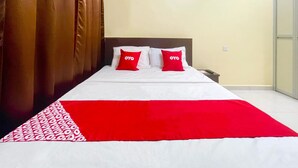 Una cama con ropa de cama blanca y roja, dos almohadas rojas con el texto "OYO", un cabecero marrón y una mesita de noche blanca.