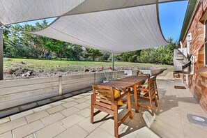 An outdoor patio with a wooden table and chairs, a covered area with a shade sail, and a brick building in the background. - Highfields - Valley Views - Bathurst Acreage (Robin Hill)