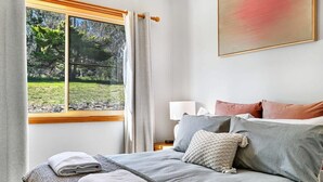 Une chambre à coucher avec un lit, des oreillers, une table de chevet, une lampe et une fenêtre avec des rideaux.