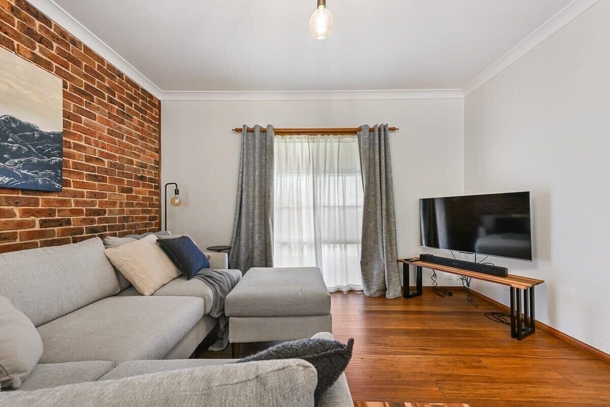 A living room with a brick accent wall, a grey sofa, a TV on a stand, and a window with curtains.
