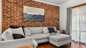 Un salon avec un mur en briques, un canapé d’angle, une petite table et une fenêtre avec des rideaux.