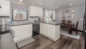 A modern kitchen with white cabinetry, a dishwasher, and a dining area with a wooden table and chairs. - Champion Center Accommodations - 6 beds, 2 bath (Appleton)